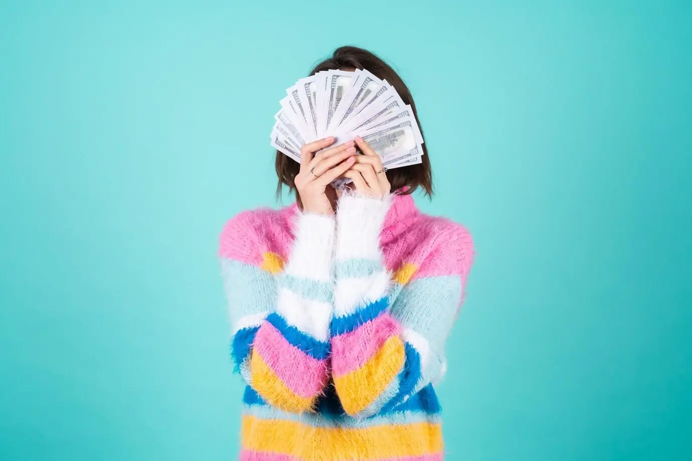Молодая женщина в ярком разноцветном свитере на синем фоне держит пачку долларов.